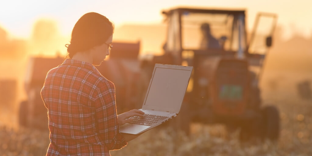 Jovem mulher com laptop em pe no campo ao por do sol enquanto o trator enfardamento no fundo representando indicadores agricolas