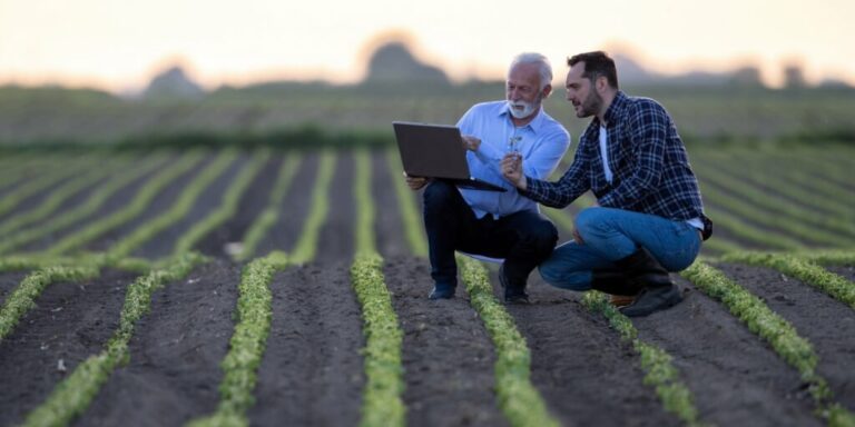 Dois homens em meio ao campo com um computador analisando as acoes do agronegocio